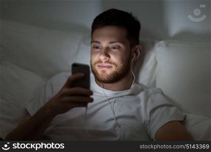technology, internet, communication and people concept - happy smiling young man with smartphone and earphones listening to music in bed at night. man with smartphone and earphones in bed at night