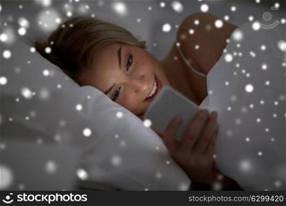 technology, internet, communication and people concept - happy smiling young woman texting on smartphone in bed at home bedroom at night over snow. young woman with smartphone in bed at home bedroom