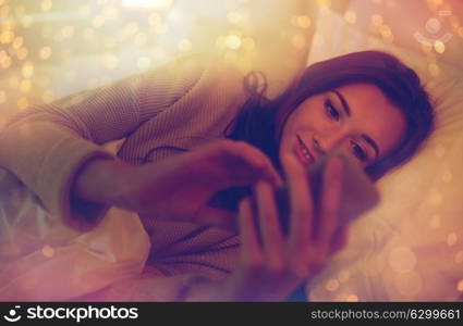 technology, internet, communication and people concept - happy smiling young woman texting on smartphone in bed at home bedroom at night. young woman with smartphone in bed at home bedroom