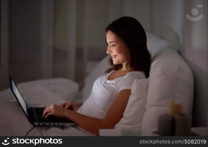 technology, internet, communication and people concept - happy smiling young woman with laptop computer in bed at home bedroom at night. happy woman with laptop in bed at home bedroom