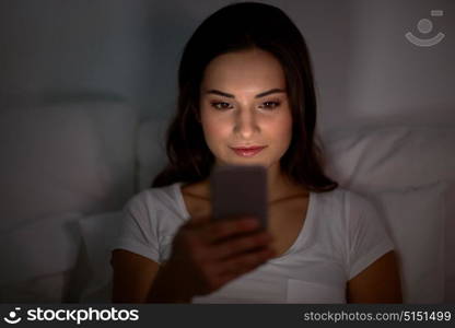 technology, internet, communication and people concept - happy smiling young woman with smartphone in bed at home at night. happy young woman with smartphone in bed at night