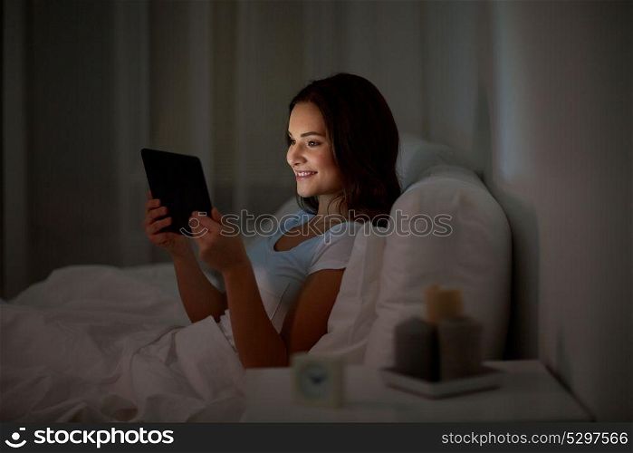 technology, internet, communication and people concept - happy smiling young woman with tablet pc computer in bed at home bedroom at night. young woman with tablet pc in bed at home bedroom