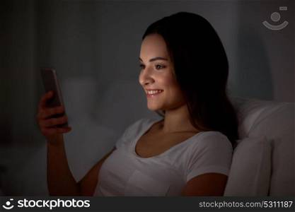 technology, internet, communication and people concept - happy smiling young woman with smartphone in bed at home at night. happy young woman with smartphone in bed at night
