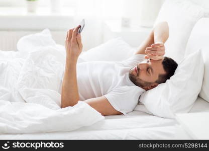 technology, internet, communication and people concept - happy young man texting on smartphone in bed at home in morning. young man with smartphone in bed in morning