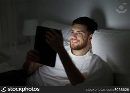 technology, internet, communication and people concept - happy young man with tablet pc computer in bed at home bedroom at night. young man with tablet pc in bed at home bedroom