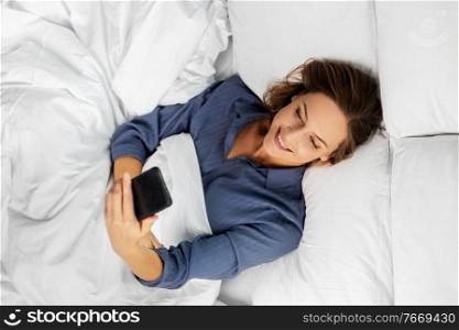 technology, internet, communication and people concept - happy young woman texting on smartphone lying in bed at home in morning. young woman with smartphone lying in bed