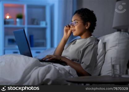 technology, internet, communication and people concept - young african american woman in glasses with laptop computer lying in bed at home at night. woman with laptop in bed at home at night