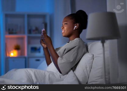 technology, internet, communication and people concept - young african american woman with smartphone and earphones listening to music in bed at home at night. woman with smartphone and earbuds in bed at night