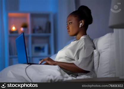 technology, internet, communication and people concept - young african american woman with laptop computer and earphones in bed at home at night. woman with laptop and earphones in bed at night