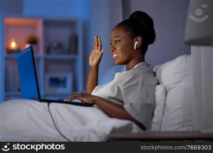 technology, internet, communication and people concept - young african american woman with laptop computer and earphones having video call in bed at home at night. woman with laptop and earphones in bed at night