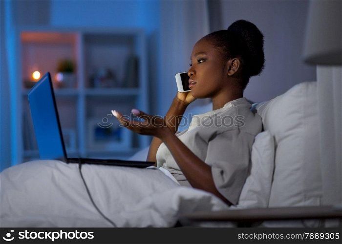 technology, internet, communication and people concept - young african american woman with laptop computer calling on smartphone in bed at home at night. woman with laptop calling on smartphone in bed