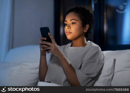 technology, internet, communication and people concept - young african american woman with smartphone lying in bed at home at night. african woman with smartphone in bed at night