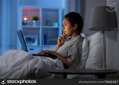 technology, internet, communication and people concept - young african american woman with laptop computer lying in bed at home at night. woman with laptop in bed at home at night
