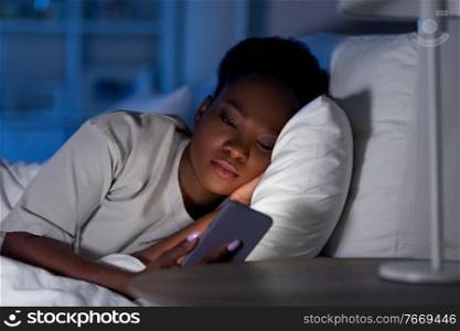 technology, internet, communication and people concept - young african american woman with smartphone lying in bed at home at night. african woman with smartphone in bed at night