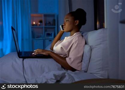 technology, internet, communication and people concept - young african american woman with laptop computer calling on smartphone in bed at home at night. woman with laptop calling on phone in bed at night
