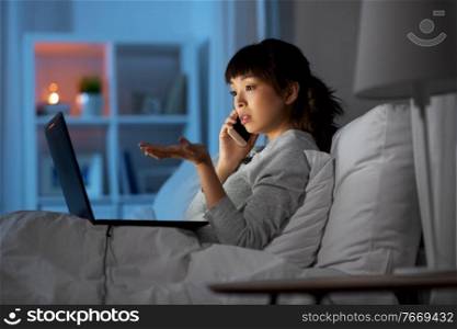 technology, internet, communication and people concept - young asian woman with laptop computer calling on smartphone in bed at home at night. woman with laptop calling on phone in bed at night