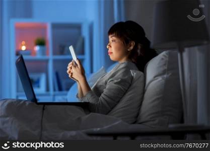 technology, internet, communication and people concept - young asian woman with smartphone and laptop lying in bed at home at night. asian woman with smartphone in bed at night