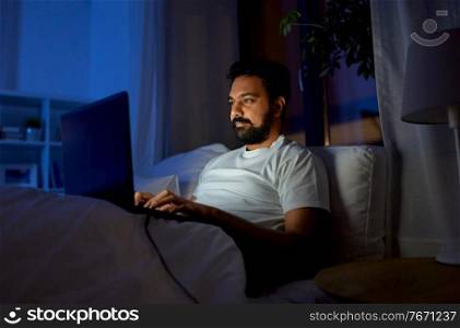 technology, internet, communication and people concept - young indian man with laptop computer lying in bed at home at night. indian man with laptop in bed at home at night