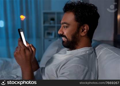 technology, internet, communication and people concept - young indian man with smartphone lying in bed at home at night. indian man with smartphone in bed at home at night