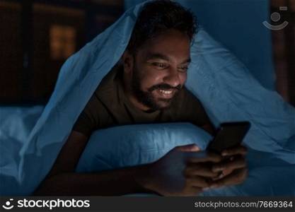 technology, internet, communication and people concept - young indian man with smartphone lying in bed under blanket at home at night. indian man with smartphone in bed at home at night