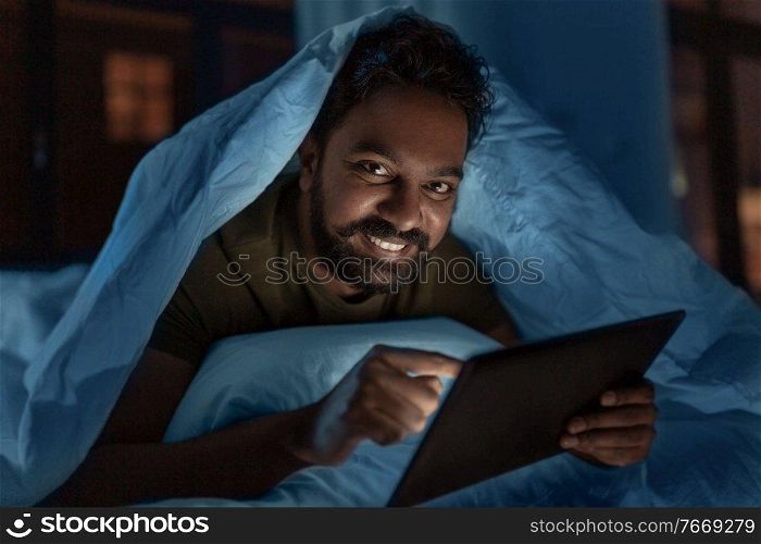 technology, internet, communication and people concept - young indian man with tablet pc computer lying in bed under blanket at home at night. indian man with tablet pc in bed at home at night