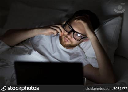 technology, internet, communication and people concept - young man in glasses with laptop computer calling on smartphone in bed at home bedroom at night. man with laptop calling on smartphone at night