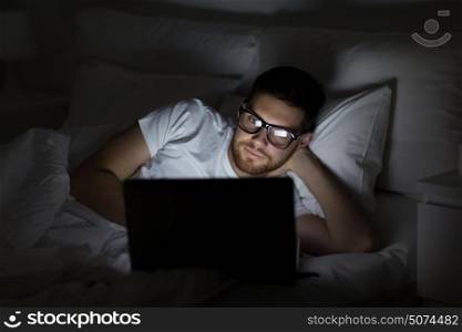 technology, internet, communication and people concept - young man in glasses with laptop computer in bed at home bedroom at night. young man with laptop in bed at home bedroom