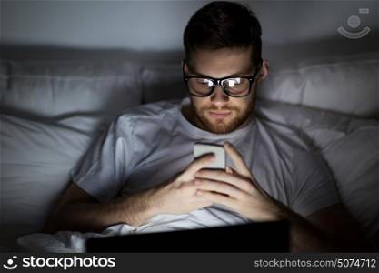technology, internet, communication and people concept - young man in glasses with laptop computer and smartphone in bed at home bedroom at night. man with laptop and smartphone at night in bed