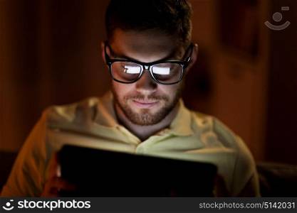 technology, internet, communication and people concept - young man in glasses with tablet pc computer networking at night at home. young man with tablet pc networking at night