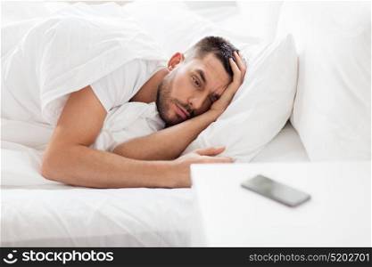 technology, internet, communication and people concept - young man lying in bed at home in morning and looking at smartphone. young man looking at smartphone