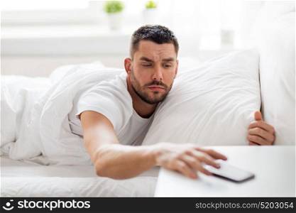 technology, internet, communication and people concept - young man reaching for smartphone in bed at home in morning. young man reaching for smartphone in bed