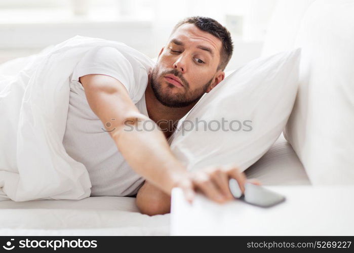 technology, internet, communication and people concept - young man reaching for smartphone in bed at home in morning. young man reaching for smartphone in bed