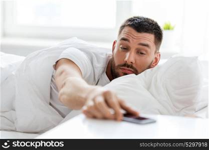technology, internet, communication and people concept - young man reaching for smartphone in bed at home in morning. young man reaching for smartphone in bed