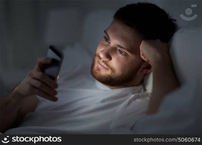 technology, internet, communication and people concept - young man texting on smartphone in bed at home at night. young man with smartphone in bed at night