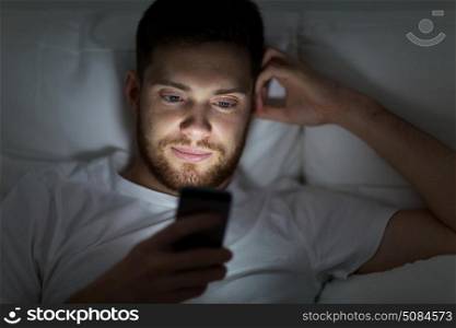 technology, internet, communication and people concept - young man texting on smartphone in bed at home at night. young man with smartphone in bed at night. young man with smartphone in bed at night
