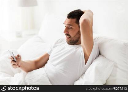technology, internet, communication and people concept - young man texting on smartphone in bed at home in morning. young man with smartphone in bed in morning