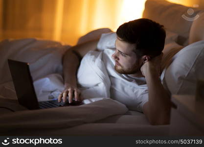 technology, internet, communication and people concept - young man with laptop computer in bed at home bedroom at night. young man with laptop in bed at home bedroom