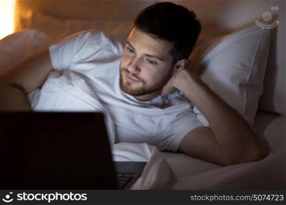 technology, internet, communication and people concept - young man with laptop computer in bed at home bedroom at night. young man with laptop in bed at home bedroom