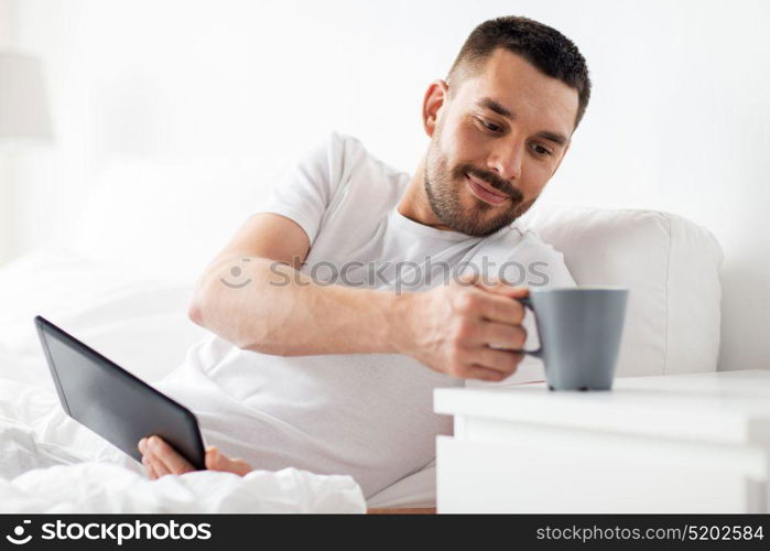 technology, internet, communication and people concept - young man with tablet pc computer drinking coffee in bed at home bedroom. man with tablet pc drinking coffee in bed at home