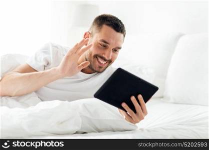 technology, internet, communication and people concept - young man with tablet pc computer having video call in bed at home bedroom. man with tablet pc having video call in bed