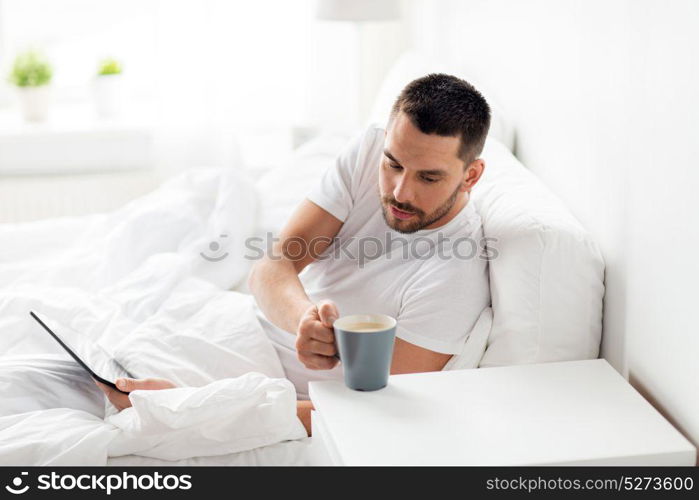 technology, internet, communication and people concept - young man with tablet pc computer drinking coffee in bed at home bedroom. man with tablet pc drinking coffee in bed at home