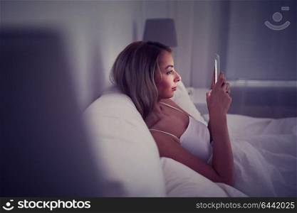 technology, internet, communication and people concept - young woman texting on smartphone in bed at home bedroom at night. young woman with smartphone in bed at home bedroom