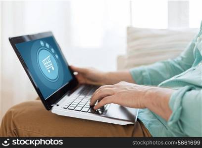 technology, leisure, advertisement and lifestyle concept - close up of man working with laptop computer displaying shopping trolley icon on screen and sitting on sofa at home