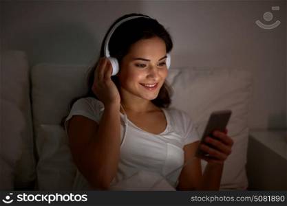 technology, leisure and people concept - happy smiling young woman with smartphone and headphones listening to music in bed at night. woman with smartphone and headphones in bed