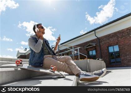 technology, leisure and people concept - indian man with smartphone, headphones and longboard listening to music on roof top. man with smartphone and headphones on roof top