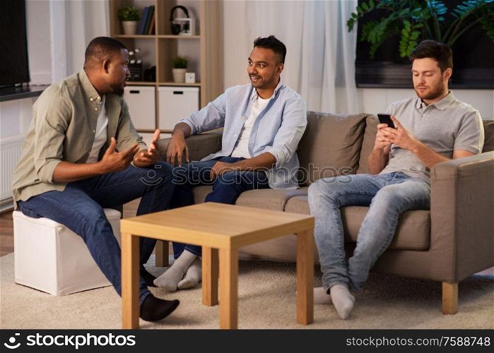 technology, leisure and people concept - man using smartphone and friends talking at home at night. man using smartphone while friends talking at home
