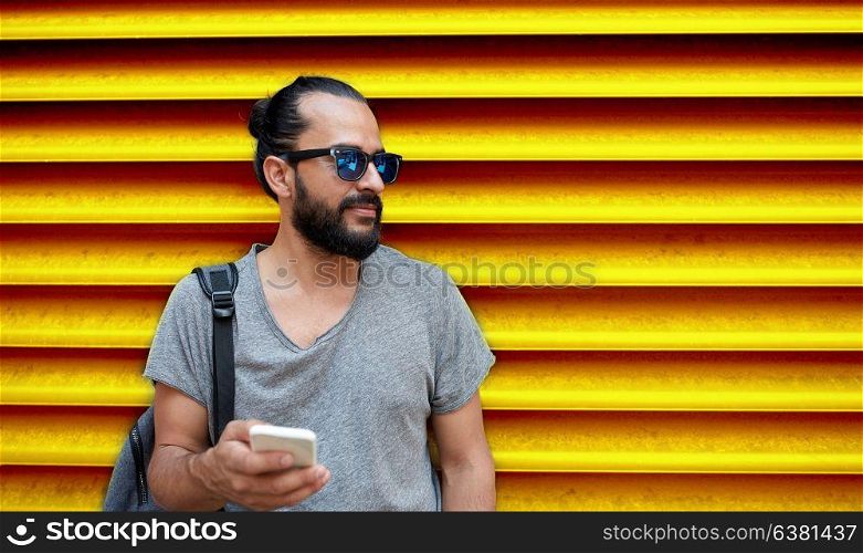 technology, lifestyle and people concept - man in sunglasses with bag and smartphone on street over ribbed yellow wall background. man in sunglasses with smartphone and bag at wall