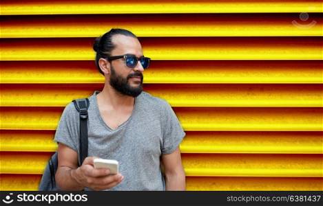 technology, lifestyle and people concept - man in sunglasses with bag and smartphone on street over ribbed yellow wall background. man in sunglasses with smartphone and bag at wall