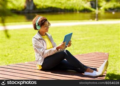 technology, lifestyle and people concept - smiling african american young woman or teenage girl with tablet pc computer and headphones listening to music or watching video in summer park