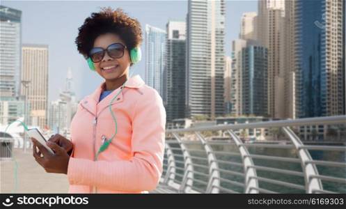 technology, lifestyle and people concept - smiling african american young woman or teenage girl with smartphone and headphones listening to music over dubai city street or waterfront background